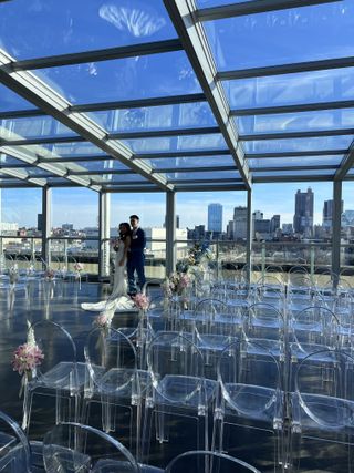 Bride and groom embracing in a glass-roofed rooftop venue with a downtown skyline view and rows of clear ghost chairs