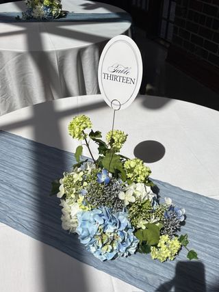 Oval “Table Thirteen” calligraphy sign above a blue-and-white hydrangea and green viburnum centerpiece on a dusty-blue runner