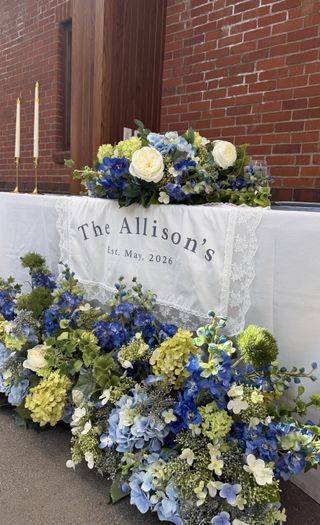 Sweetheart table draped in a lace “The Allison’s, Est. May 2026” runner banked by a lush blue, white, and green floral meadow