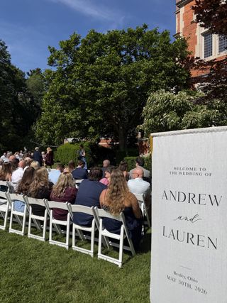 Outdoor wedding ceremony seating facing a linen “Andrew and Lauren” welcome sign beneath a large shade tree