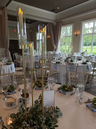 Reception ballroom set with gold chiavari chairs, tall taper candlesticks, and dusty-blue napkins overlooking a golf course
