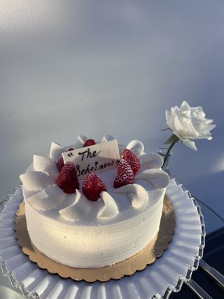 Strawberry-topped whipped-cream cake lettered “The Scheiners” beside a single white rose in a bud vase