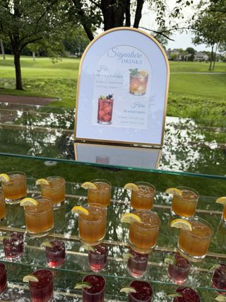 “Signature Drinks” arched menu sign beside rows of garnished cocktails on a mirrored tray with a green lawn behind