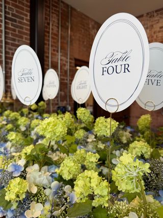 Row of oval calligraphy table-number signs rising from a bed of green viburnum and blue hydrangea against exposed brick
